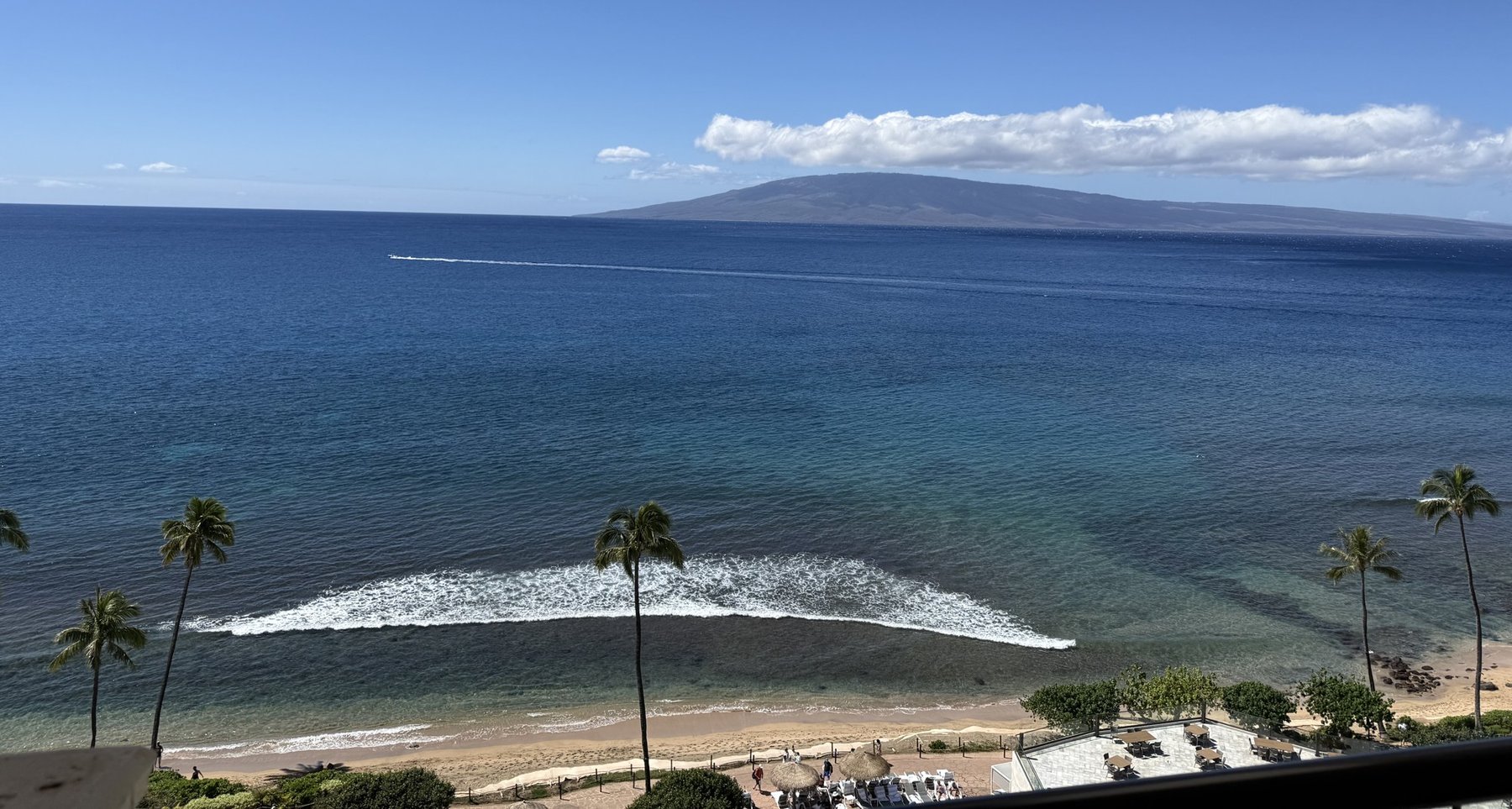 Looking across the water from Maui toward the island of Lānaʻi, with palms and a breaking wave in the foreground